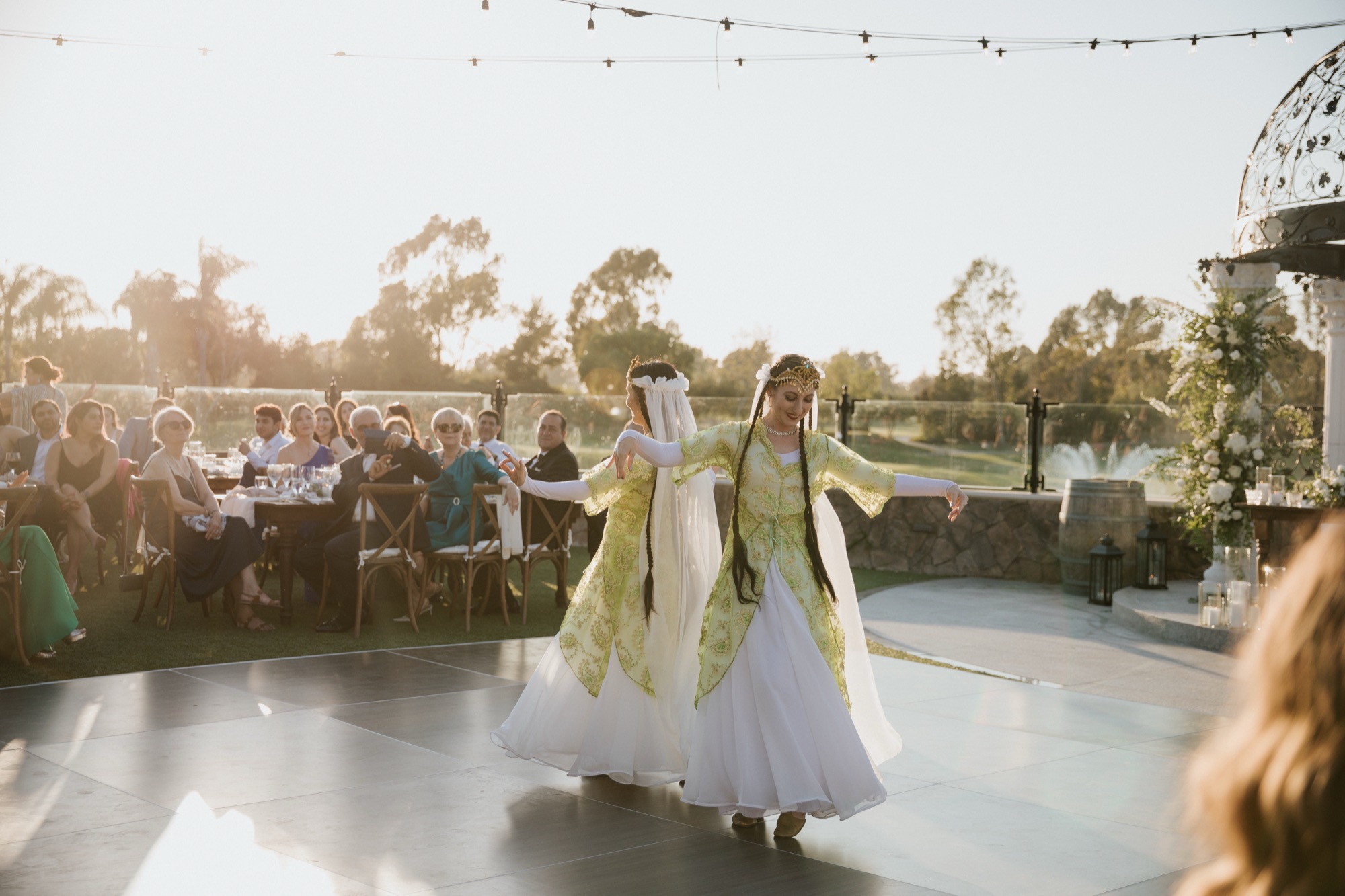 Two dancers in green and white Persian costumes performing for wedding guests at sunset