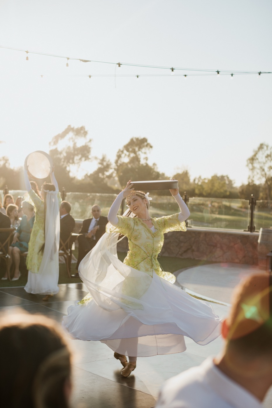 A dancer twirling in a flowing white skirt while balancing a daf drum, with wedding guests watching